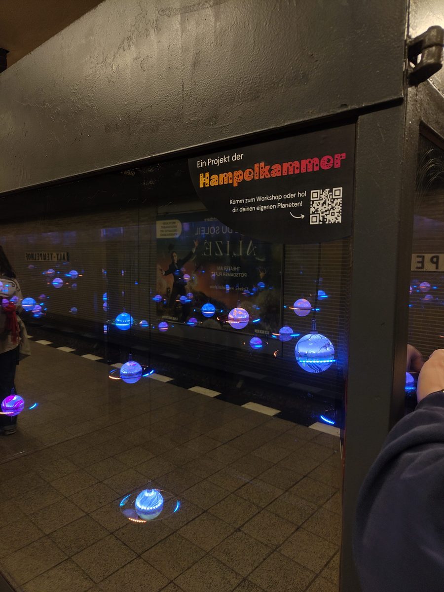 Hampelkammer-Beschilderung mit QR-Code am U-Bahnhof Alt-Tempelhof, im Hintergrund die leuchtende Planeten-Installation
